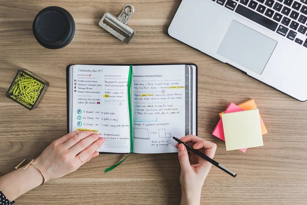 Hands writing a plan in a notebook beside a laptop